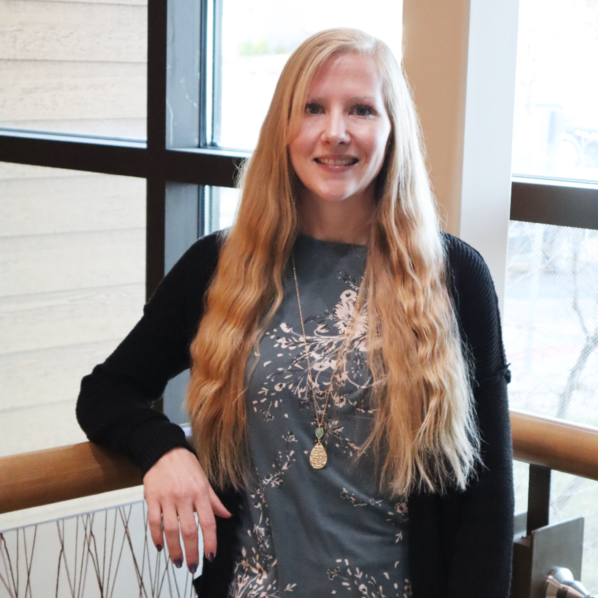 Woman smiling, looking at the camera. She has long blonde hair, wearing a light pink blouse, and a light blue necklace.
