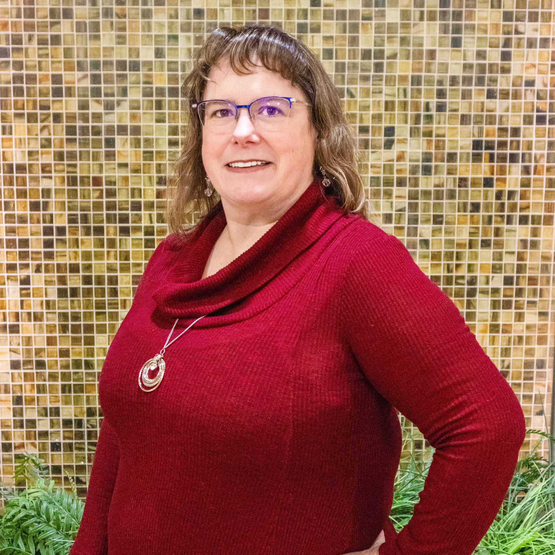 Woman with glasses smiles; wearing a blue floral top, black cardigan, and blue necklace. Indoors, near a window.