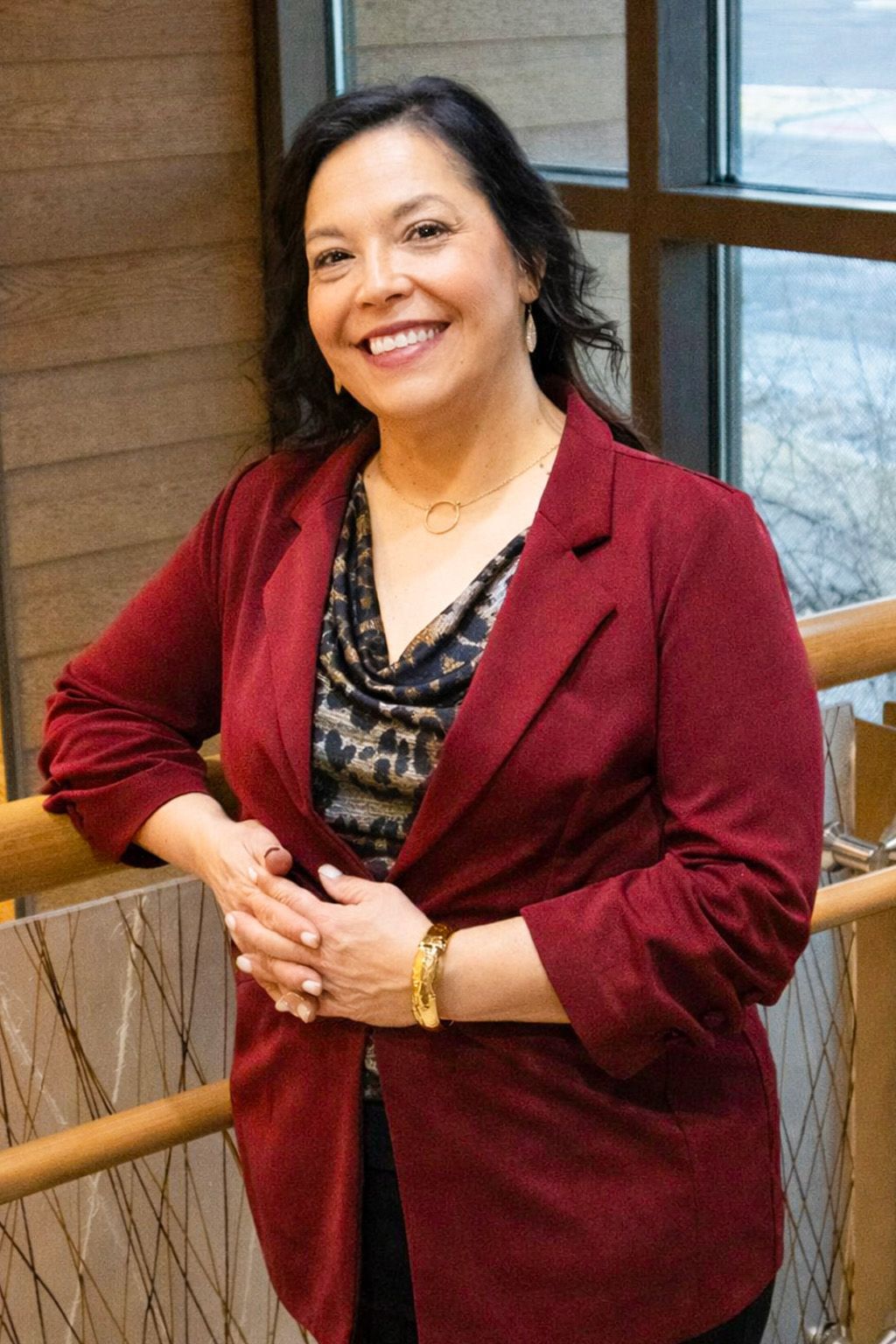 Woman smiling, black hair, wearing black top, black jacket, and hoop earrings, standing indoors.
