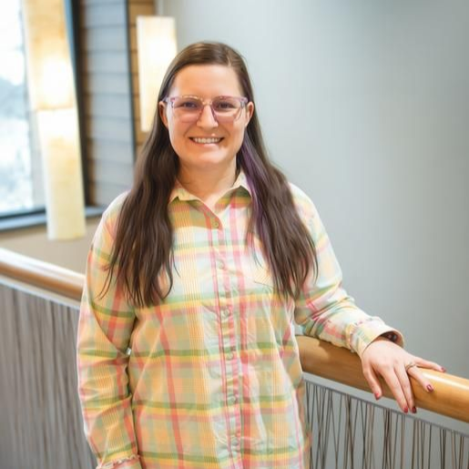 Woman with glasses smiling, wearing a black jacket and top, standing near a window.