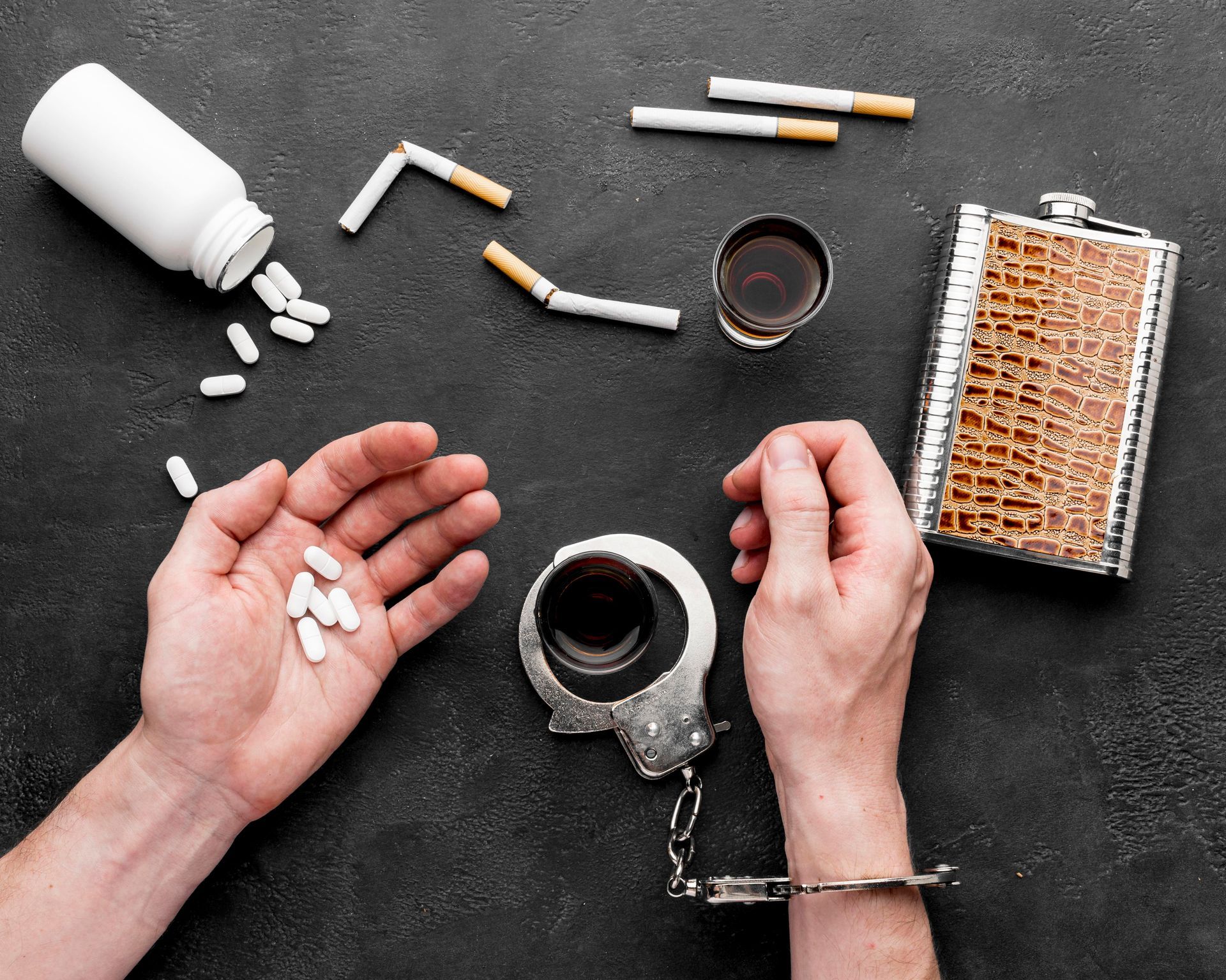 Hands with pills, cigarettes, and alcohol, one hand cuffed to a drink, depicting addiction.