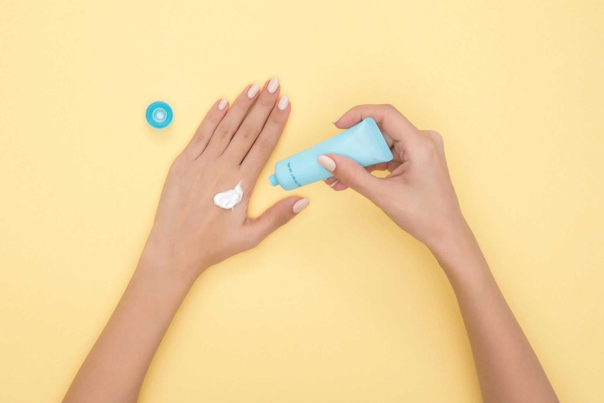 Hands squeezing white lotion from a blue tube onto the back of a hand; a blue cap sits nearby on a yellow surface.