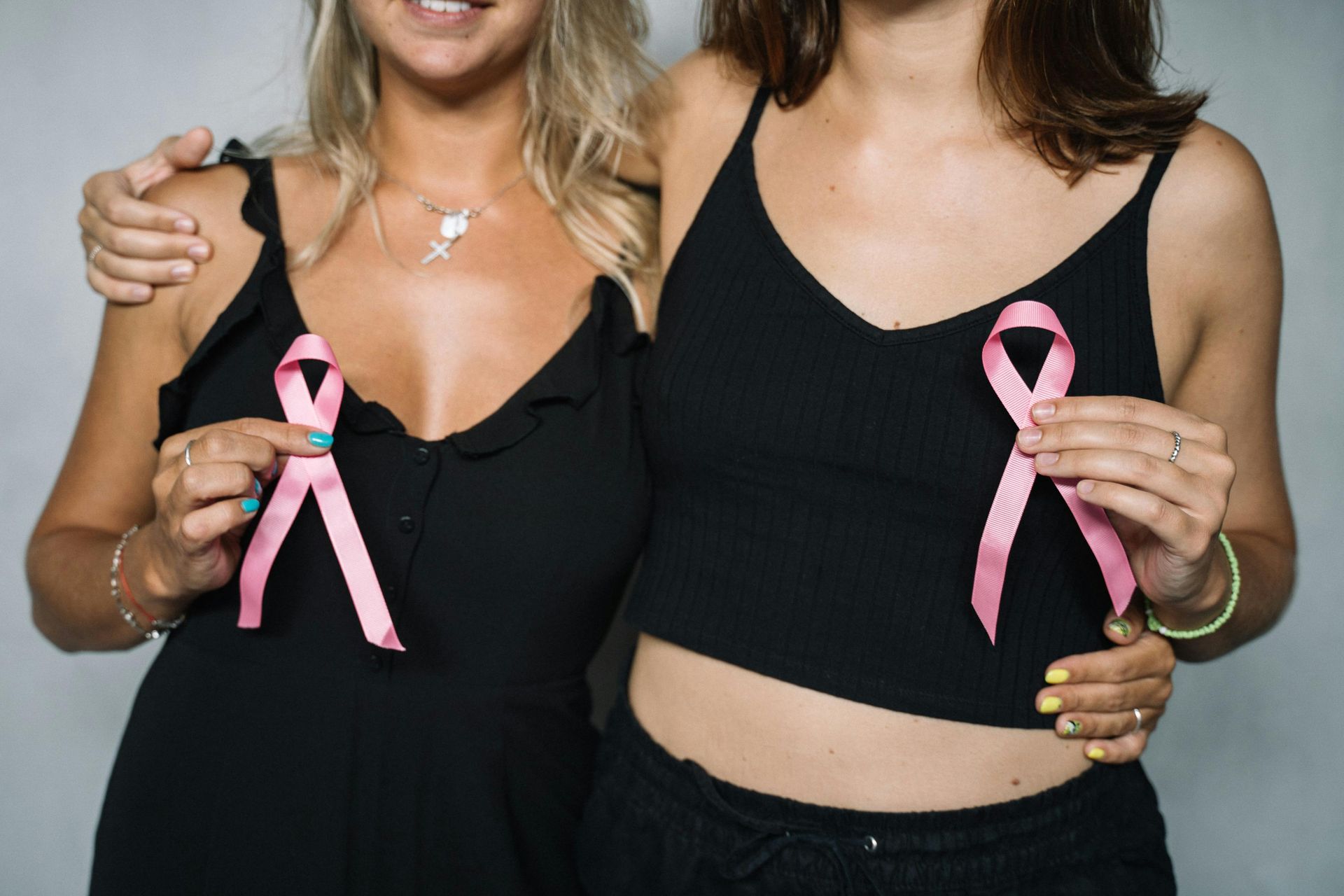 Two women holding pink ribbons, likely for breast cancer awareness, wearing black tops.