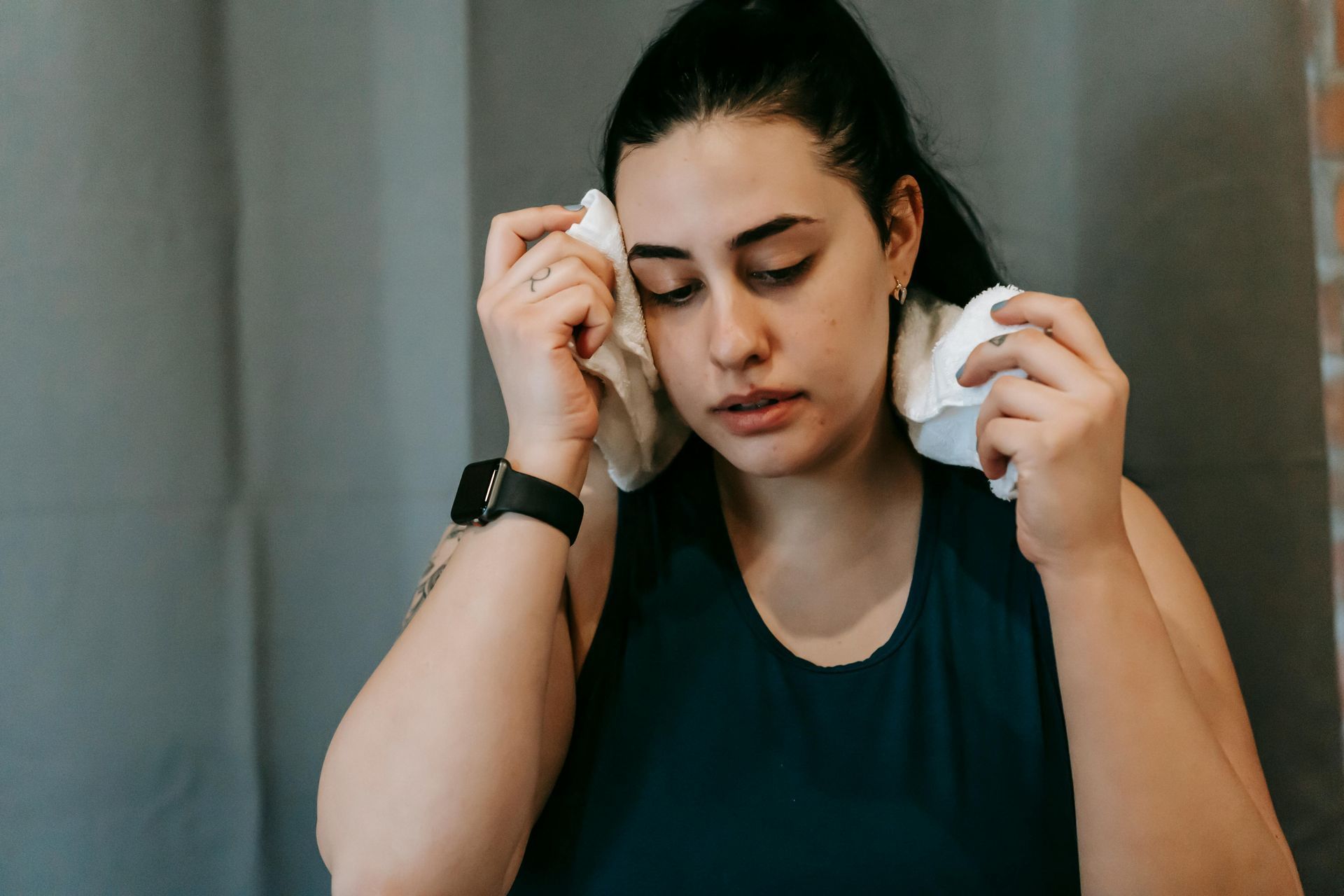 Woman wiping sweat from her face with a white towel after a workout; wearing a black smartwatch and blue tank top.