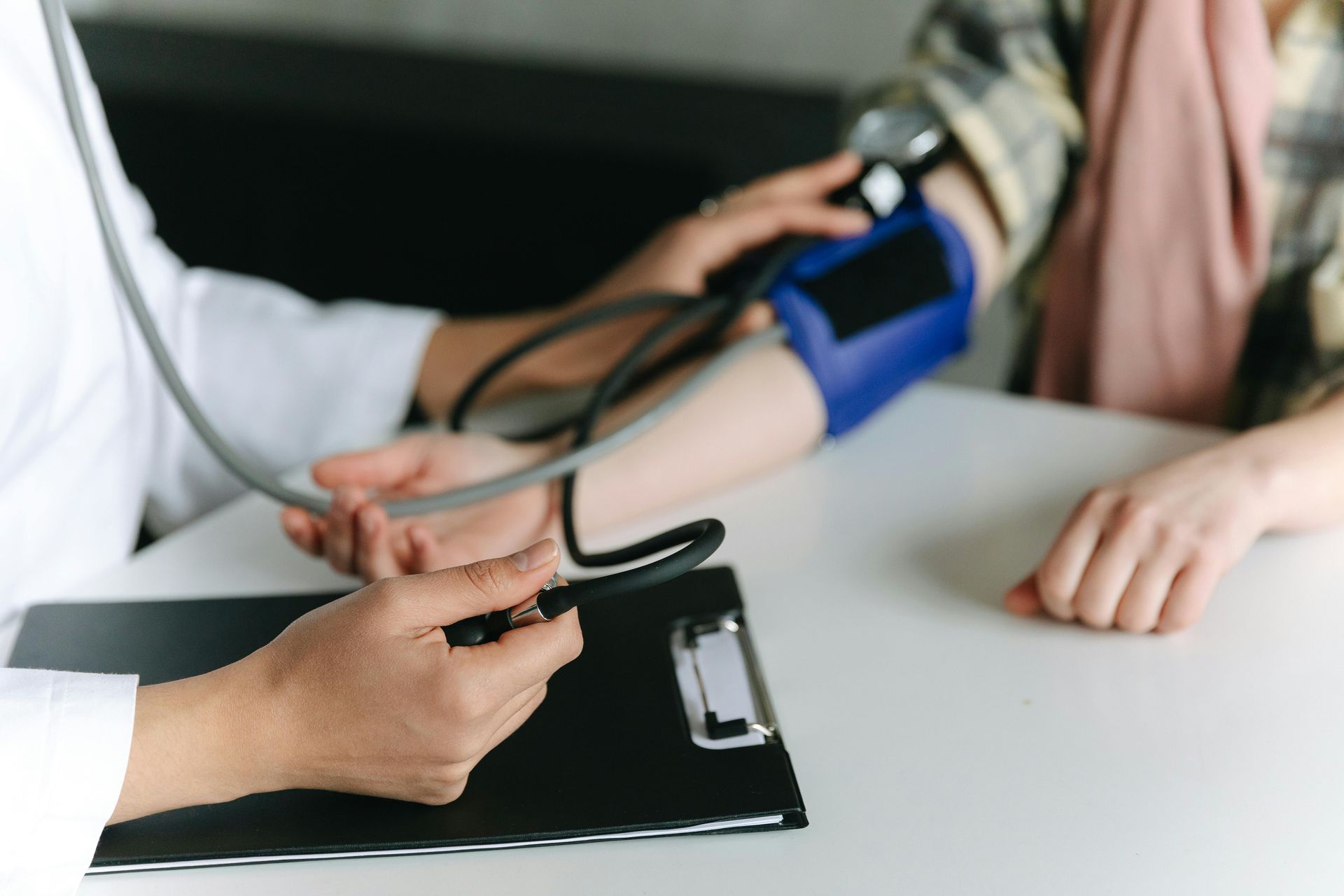 Doctor taking patient's blood pressure with stethoscope and cuff on a white table.