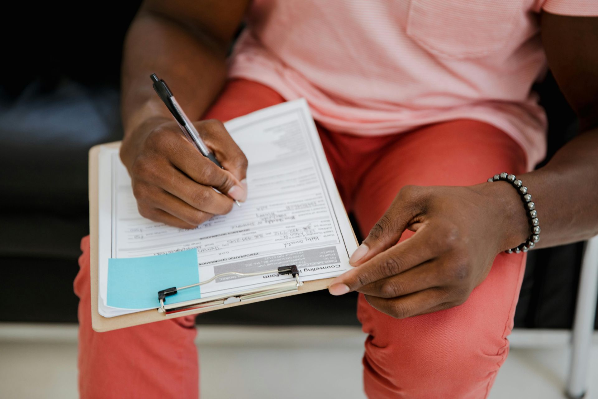 Person writing on a clipboard, wearing pink shirt and red pants.