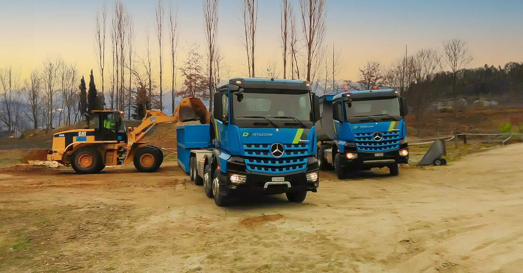 Due camion blu sono parcheggiati uno accanto all'altro con un'escavatore che li carica su una strada sterrata.