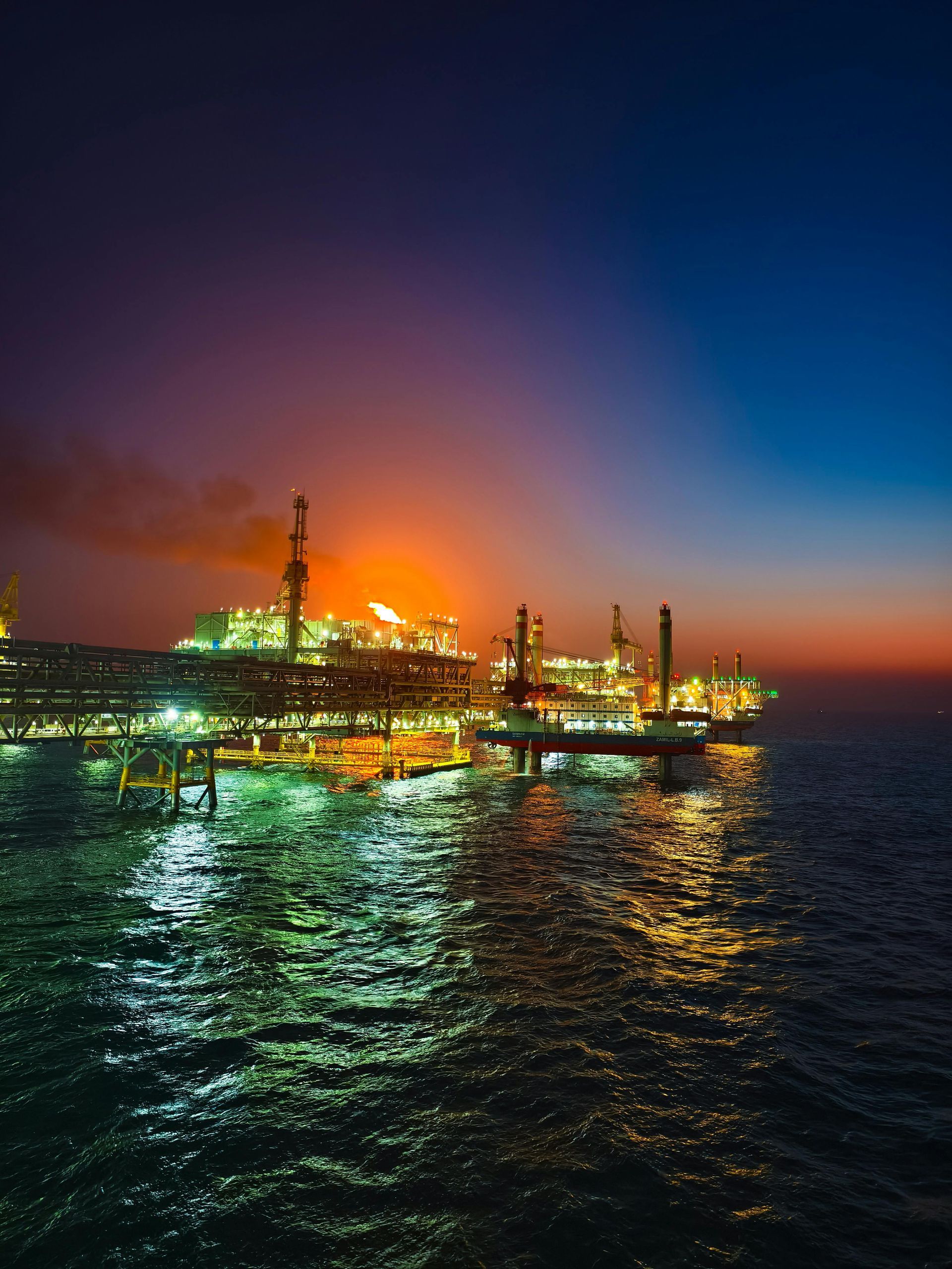 An offshore oil rig illuminated at dusk, with industrial lights reflecting on the dark, rippling ocean water.