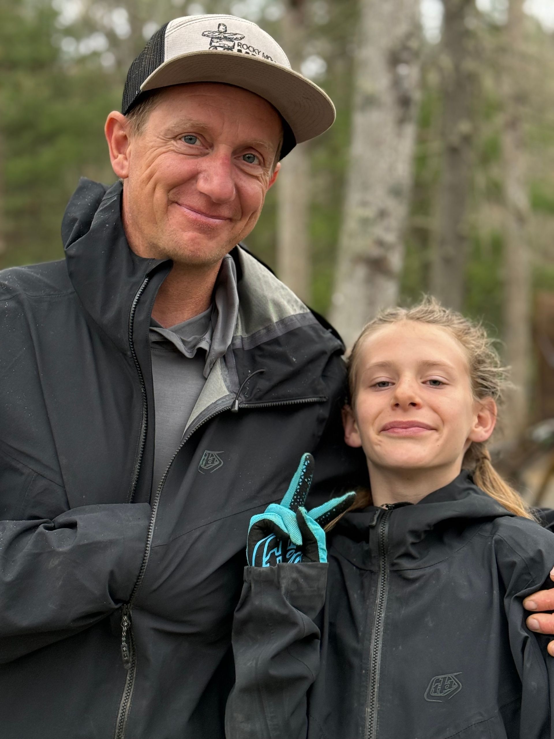Man in cap and girl in jackets, smiling outdoors. Girl makes peace sign.
