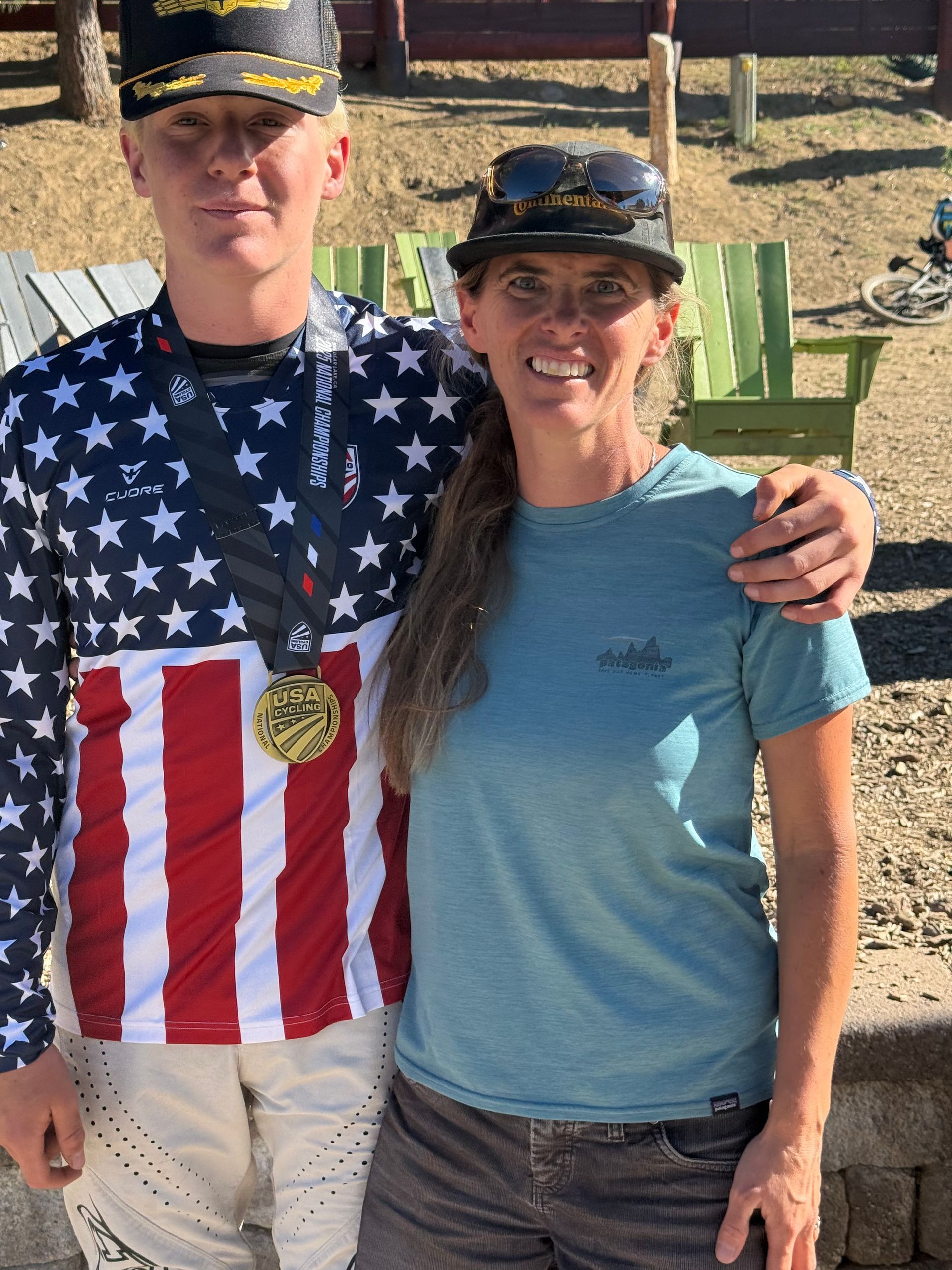 Man with medal and woman smiling, arm around each other. Outdoor setting. American flag shirt.