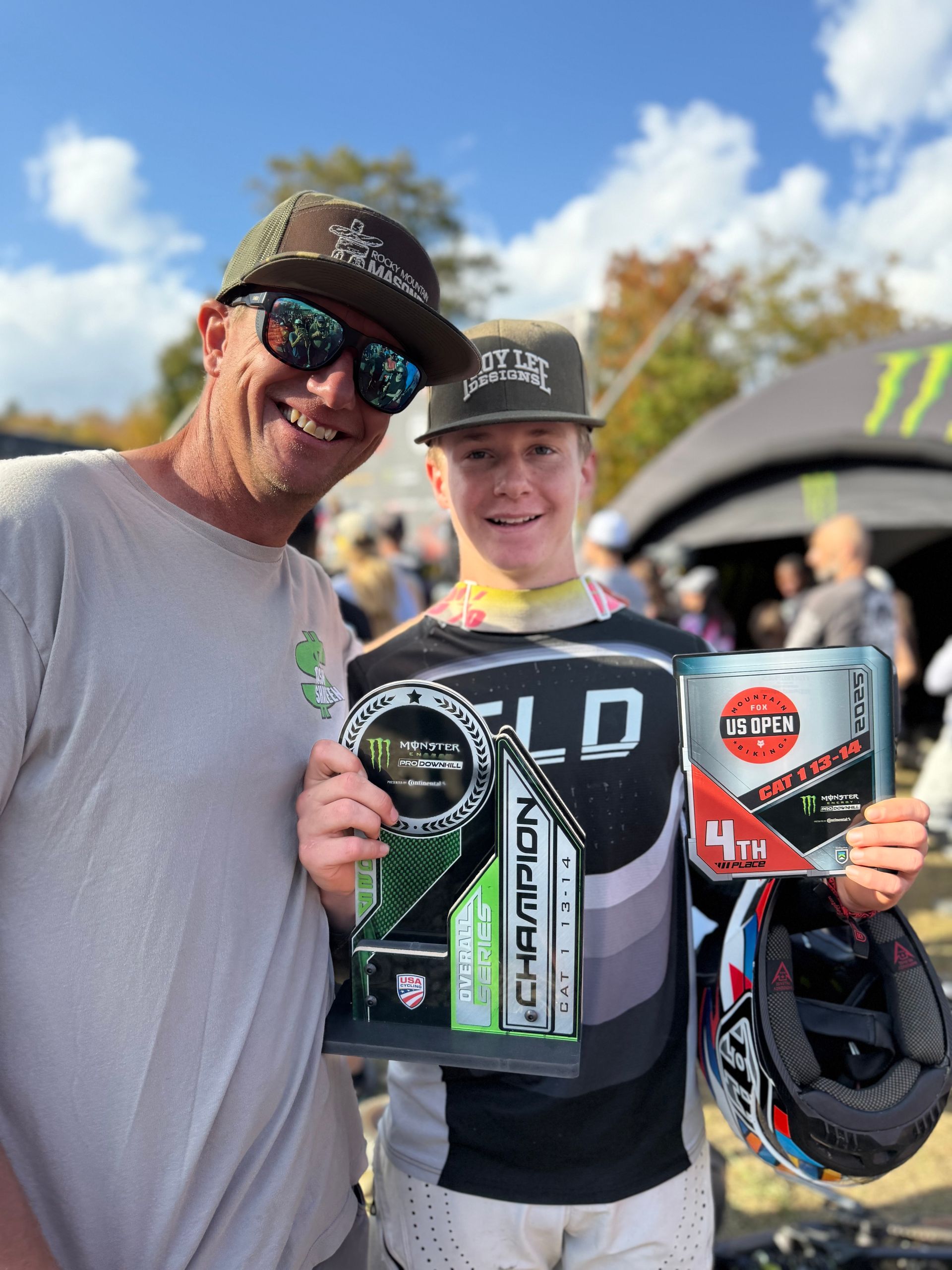 Man and young person smiling, holding a trophy and number plate, outdoors at event.
