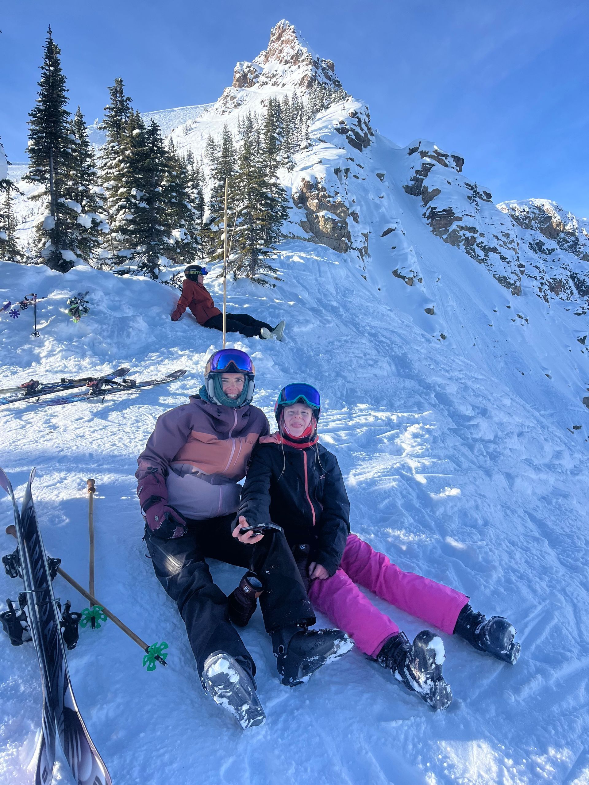 Two people in ski gear sit in snow, mountain peak in background. Person rests nearby.