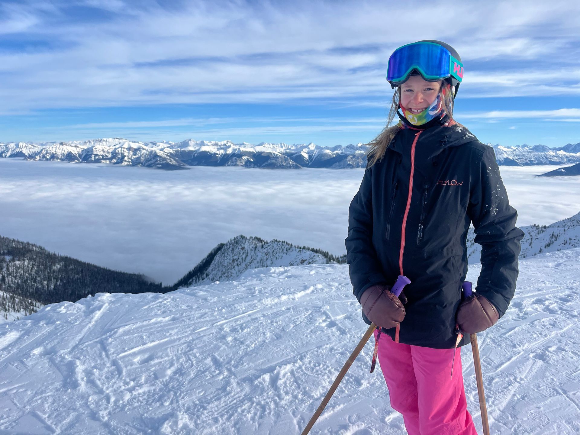 Girl in ski gear stands on snowy mountain, blue goggles, pink pants, looking at camera.