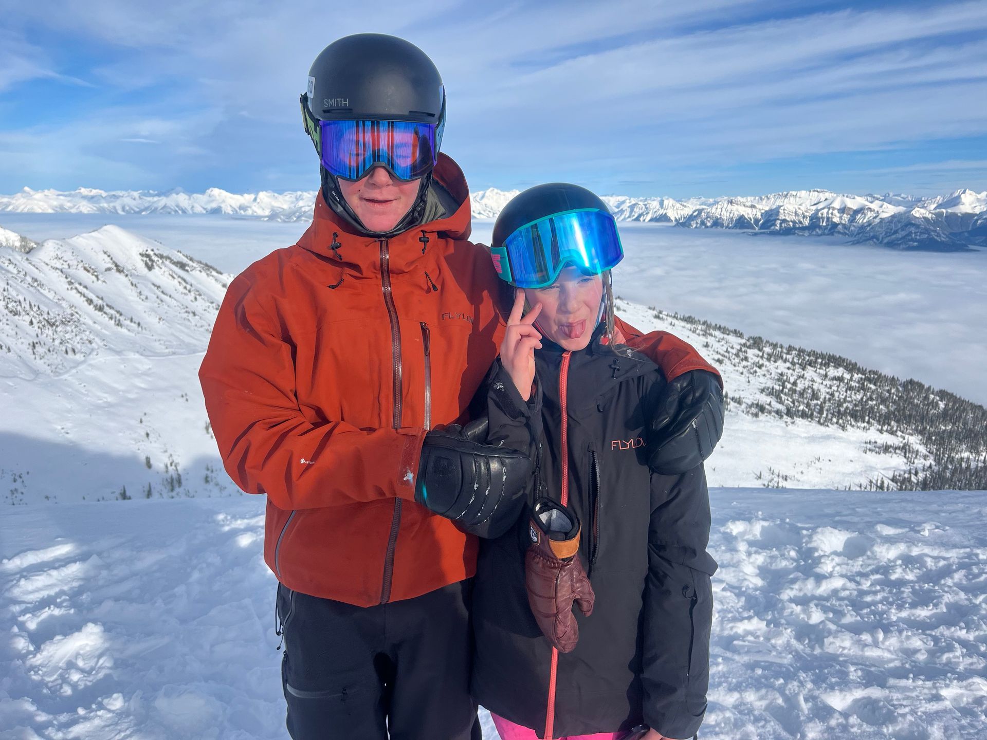 Two people in ski gear pose on a snowy mountain, with a cloudy valley in the background.