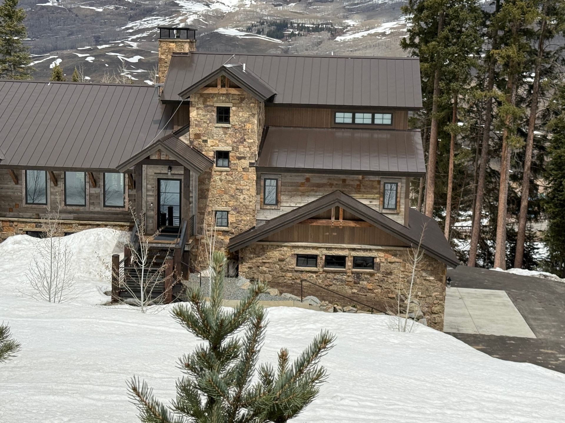 Stone and wood cabin in a snowy mountain setting, dark metal roof, trees in foreground.