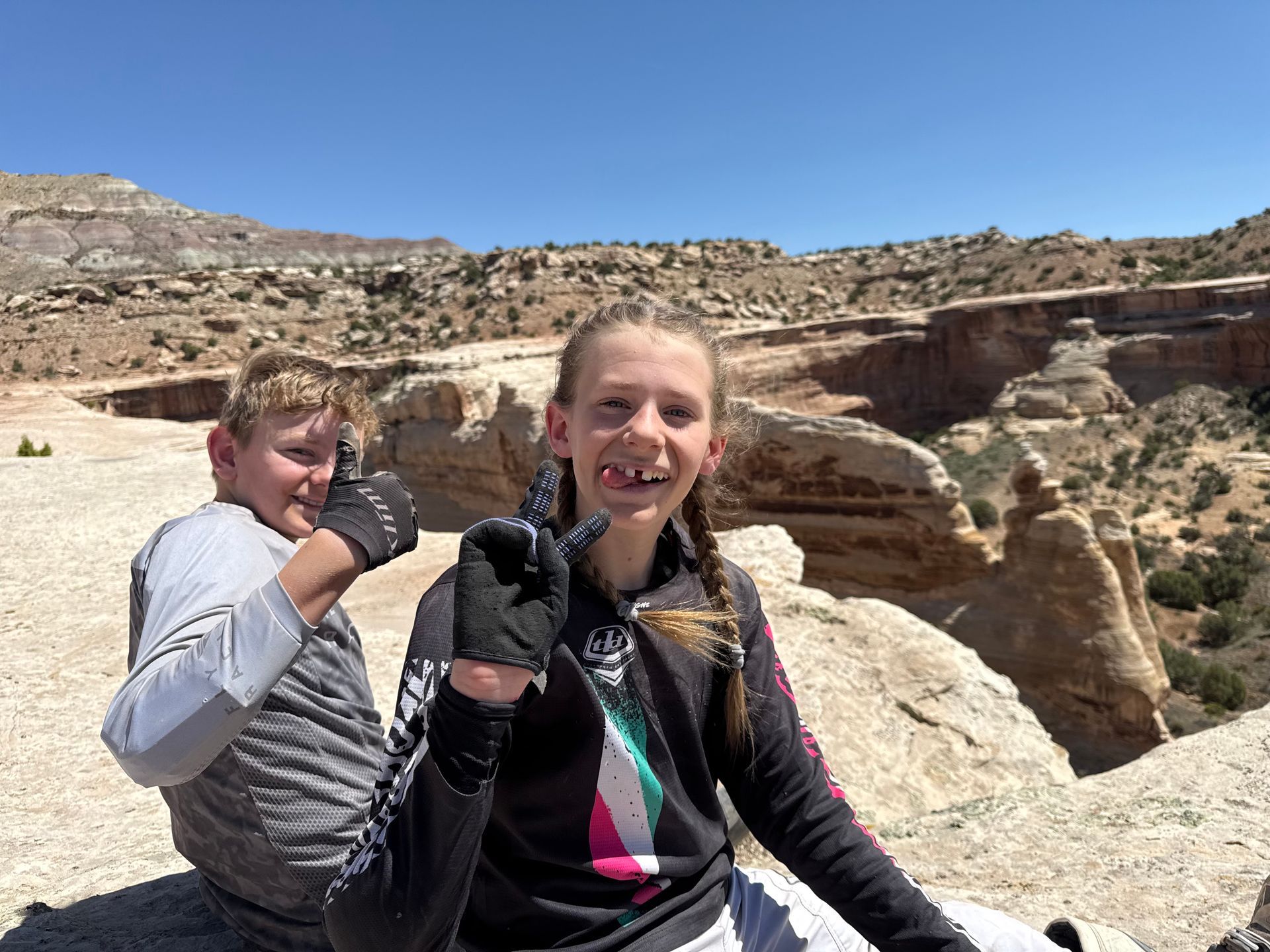 Two young people give thumbs-up, smiling outdoors on a rocky outcrop. Beige cliffs and blue sky.