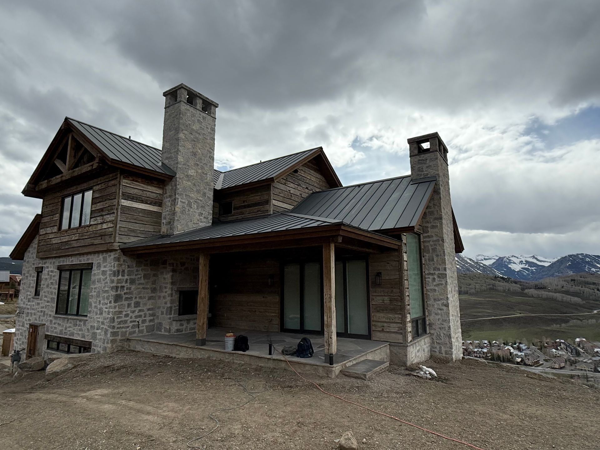 Two-story stone and wood house with chimneys, porch, and metal roof under cloudy sky.