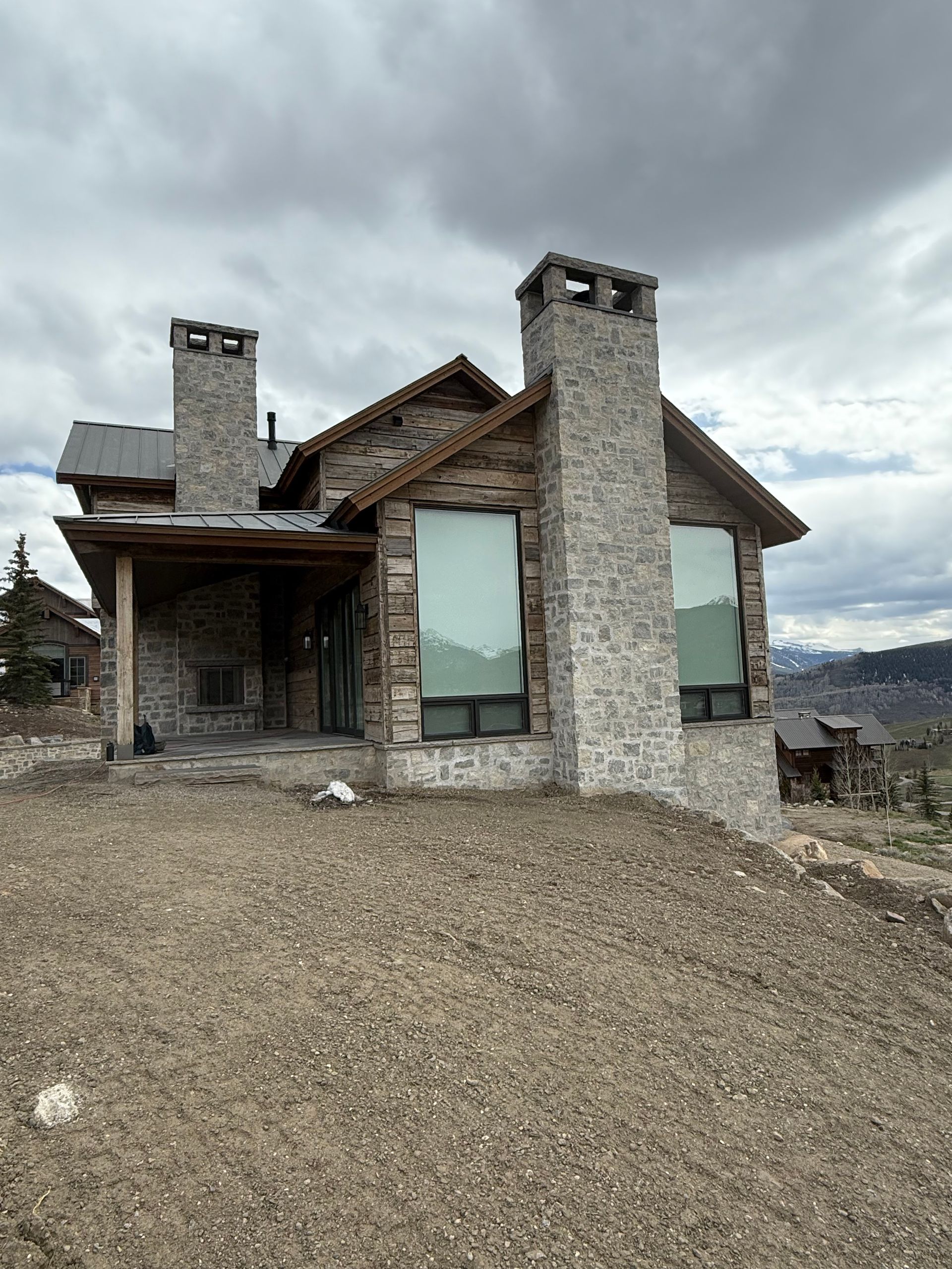 Stone house with large windows, chimneys, and a covered porch on a hillside under a cloudy sky.
