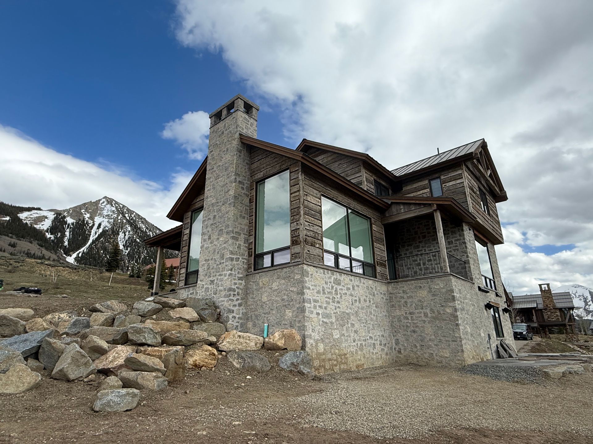 Stone house with large windows and chimney against mountain and cloudy sky.