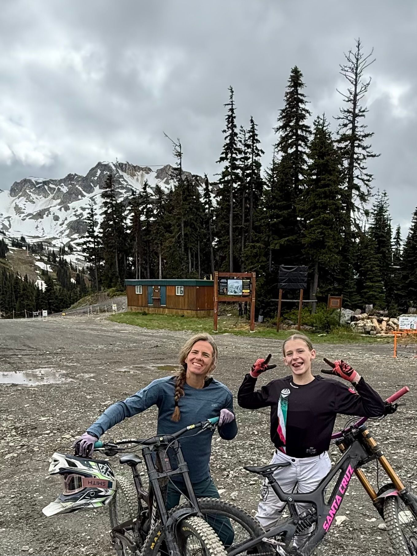 Two women with mountain bikes pose in front of a mountain and trees on a cloudy day. One makes a peace sign.