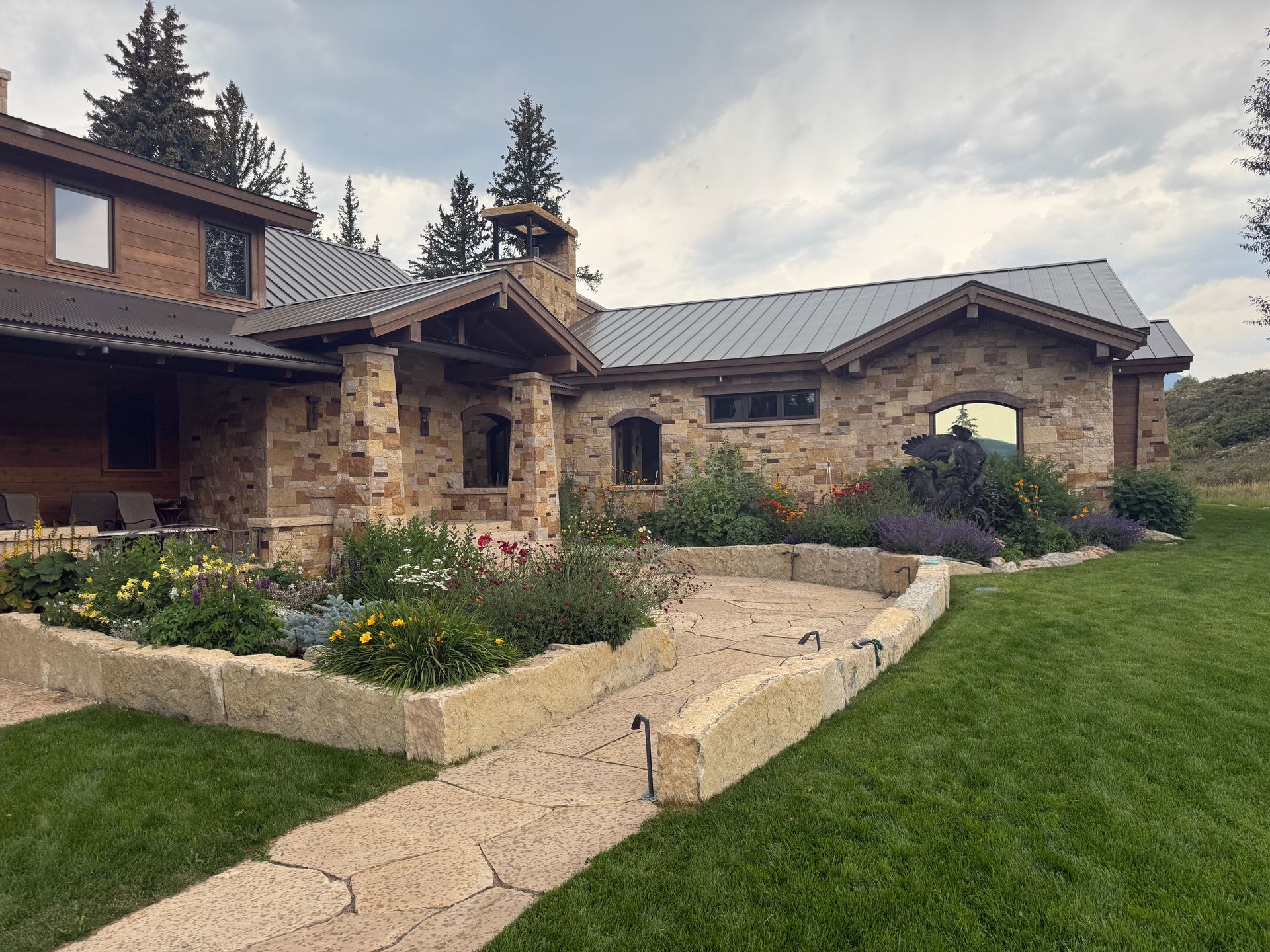 Stone house with a metal roof, a porch, and a garden bed against a grassy lawn.