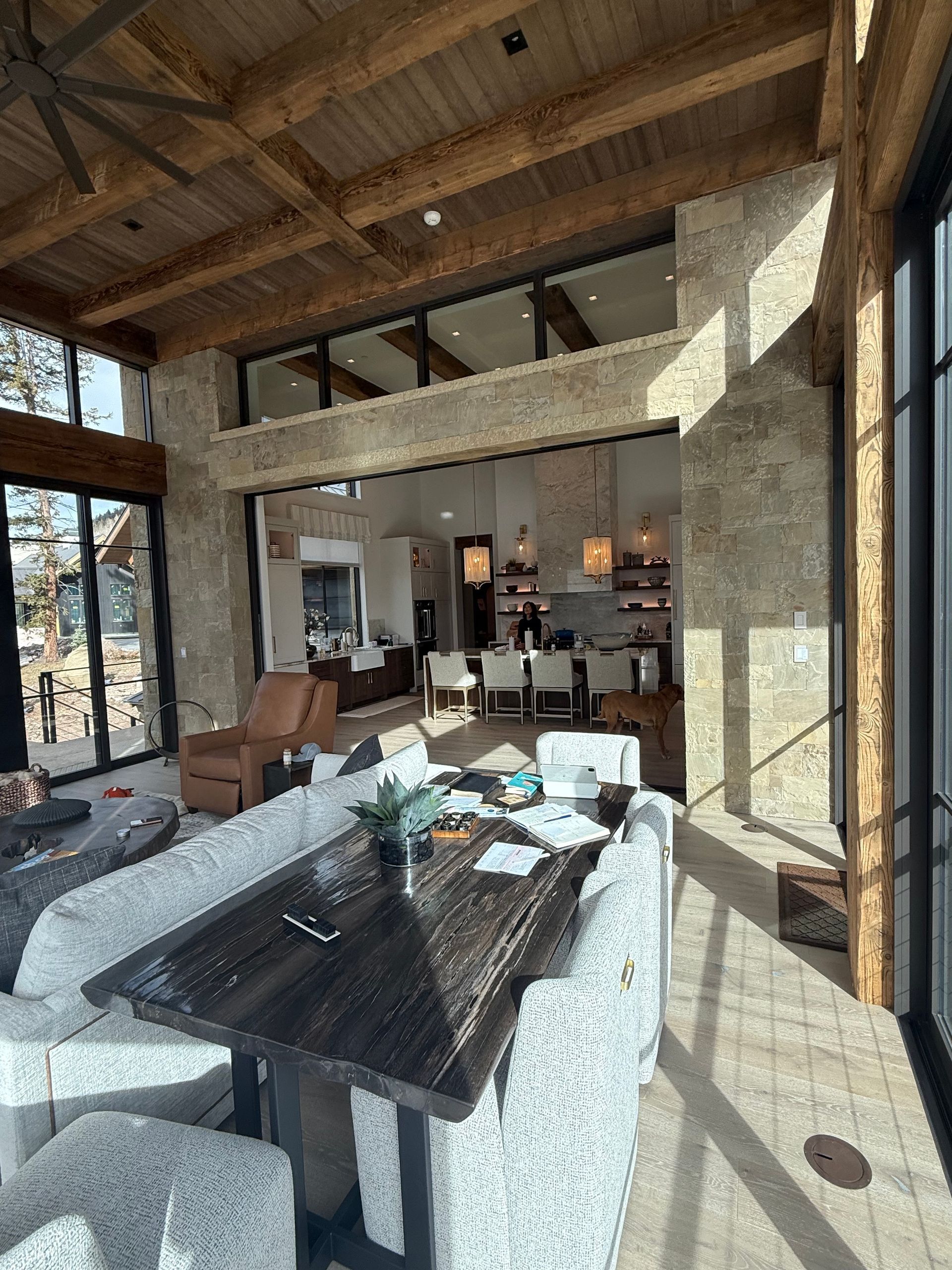 Living room with wood beams, stone wall, and dark wood table. Open to a kitchen area.