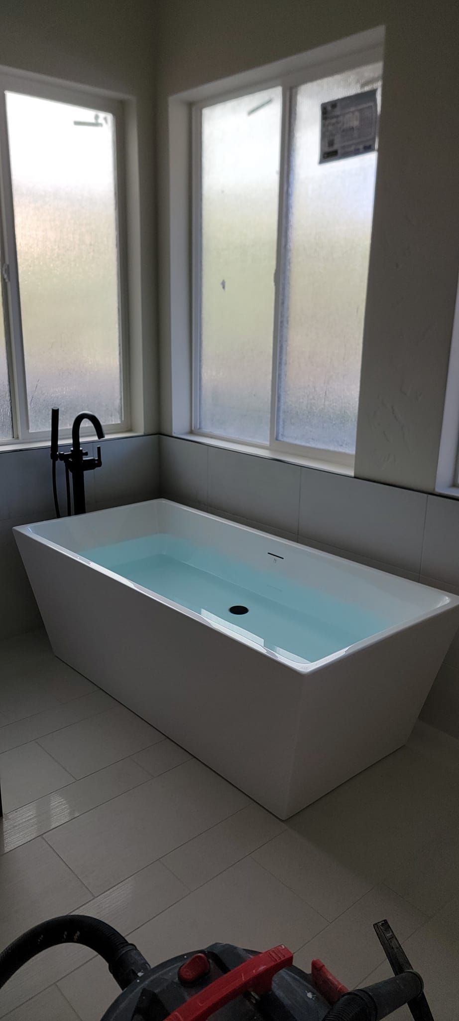 A white freestanding bathtub filled with water sits in a corner bathroom with two windows and a black faucet.