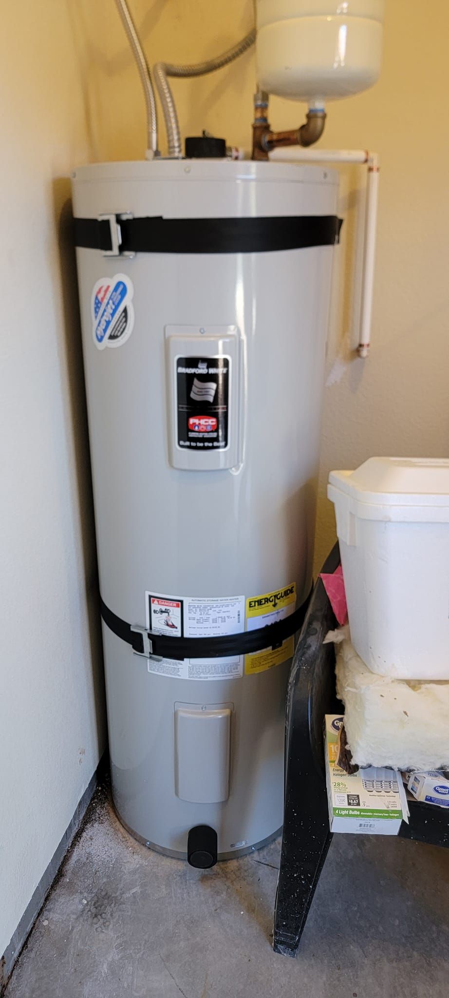A tall, light-gray cylindrical water heater with black seismic straps installed in a garage setting.