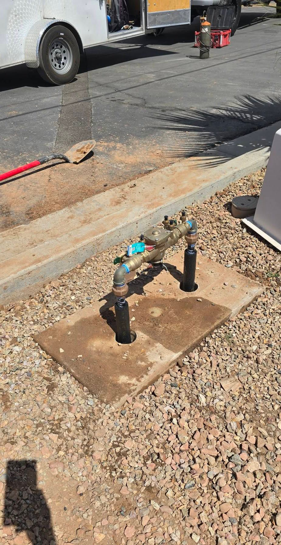 A residential water meter assembly with pipes and a brass valve, mounted on a concrete pad in a gravel landscaping area.