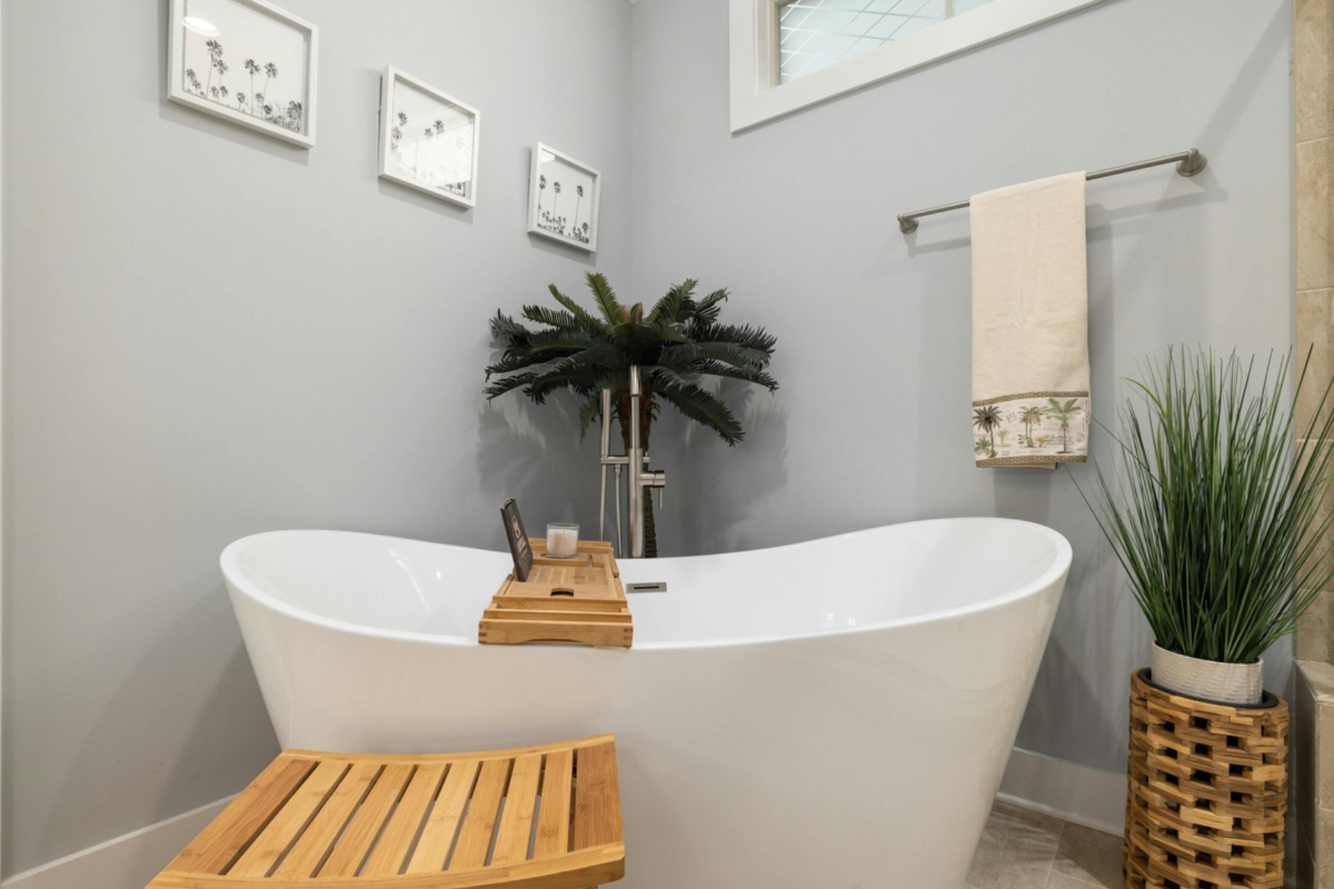 A white freestanding bathtub with a wooden tray and seat in a light grey bathroom with framed art and potted plants.