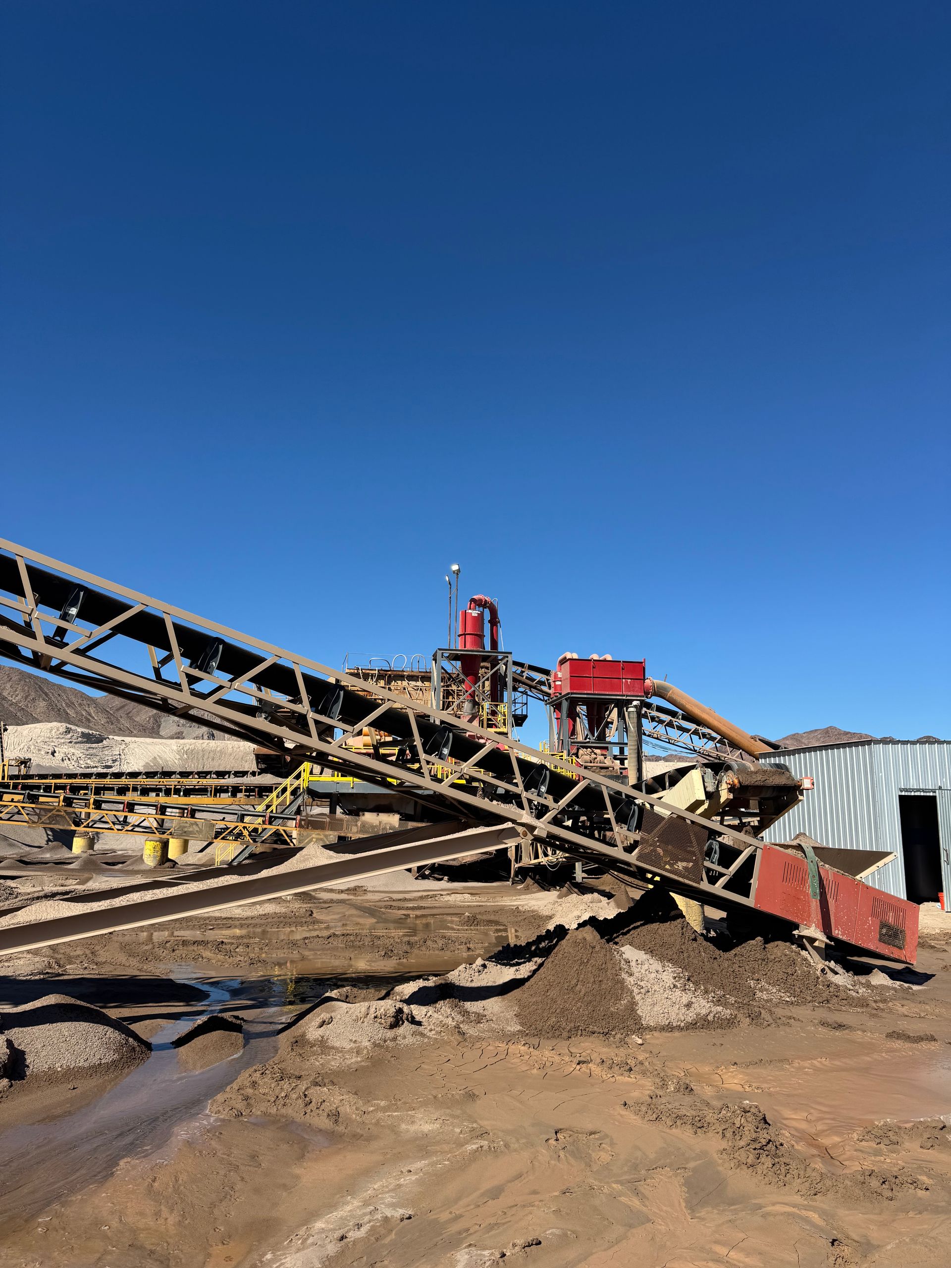 A mining operation with conveyor belts, machinery, and a red structure under a clear blue sky.