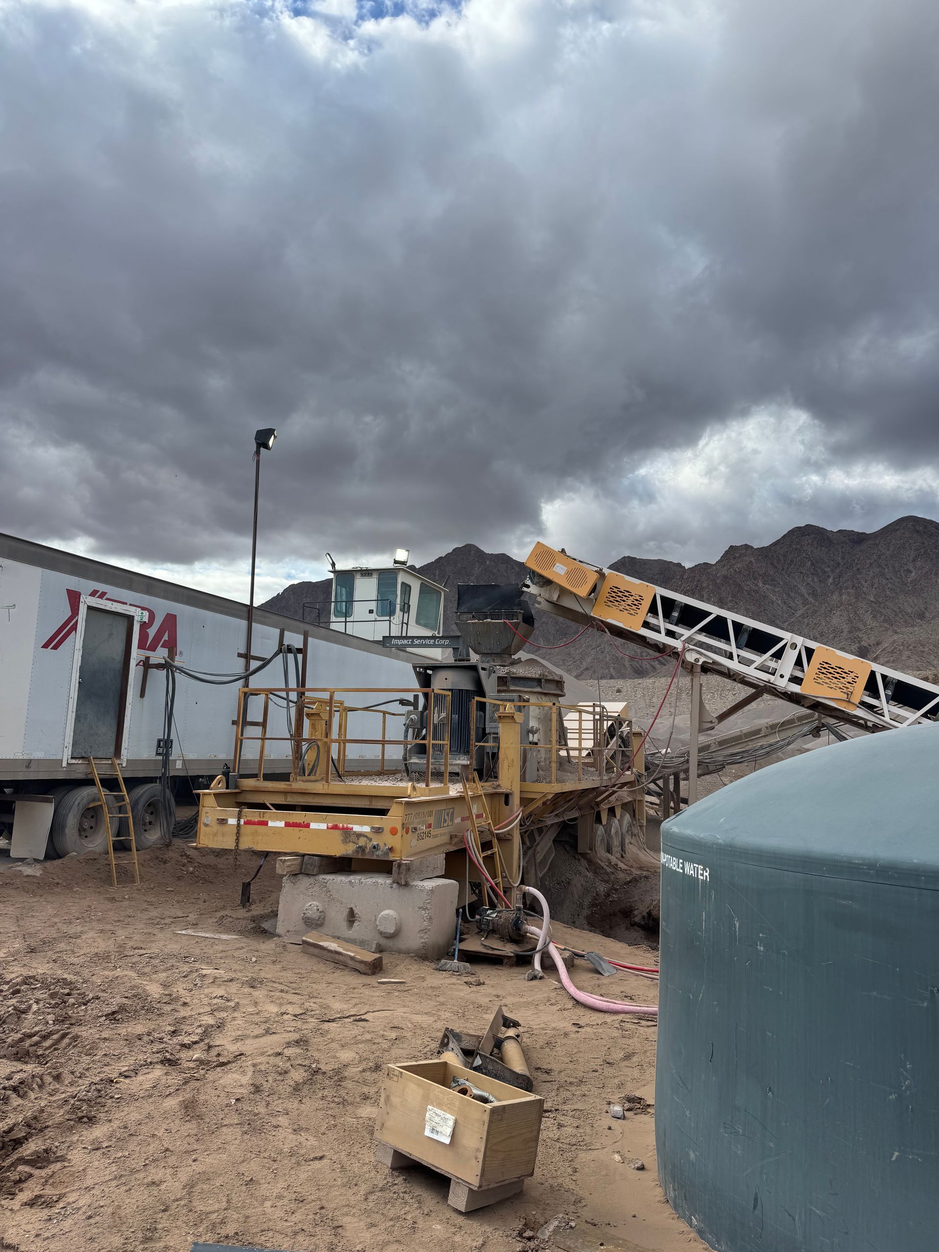 Yellow mining equipment, conveyor belt, and water tank in a desert landscape under a cloudy sky.