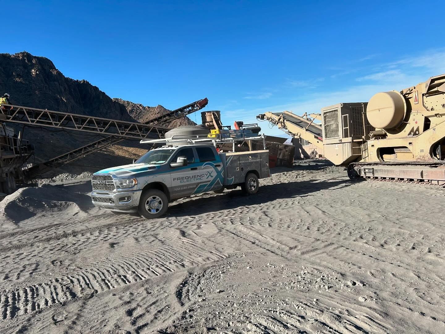A work truck parked near rock crushing equipment in a quarry, under a clear blue sky.