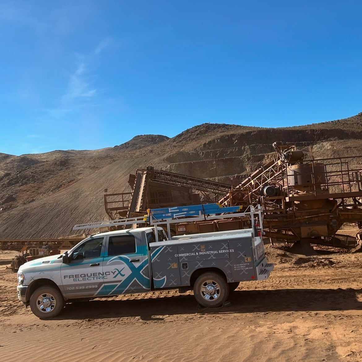 White service truck with logo parked in front of a rusty mining structure in a desert landscape.