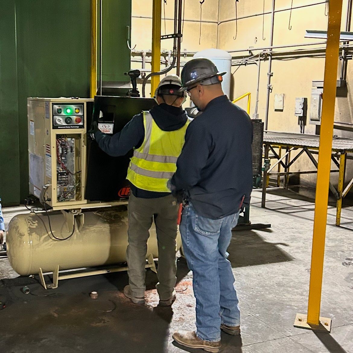 Two workers inspecting machinery in a factory. One wears a reflective vest and hard hat.