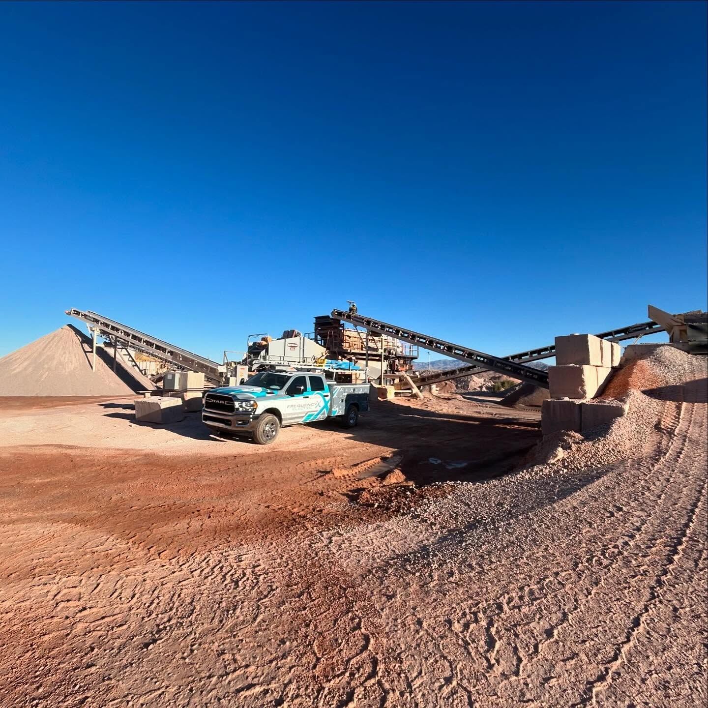 Truck at a quarry, with machinery and piles of gravel, against a clear blue sky.