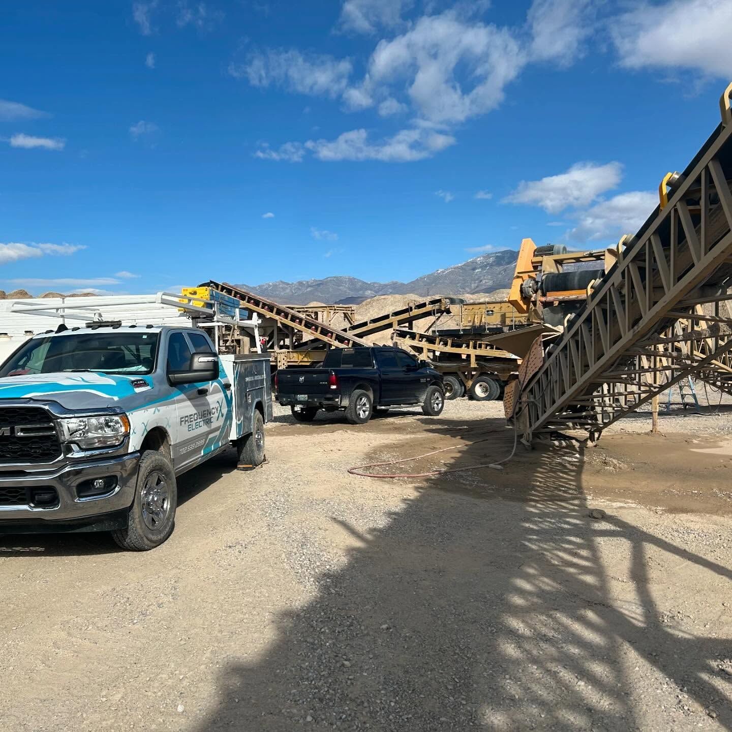 A work truck, black truck, and machinery sit in a quarry under a blue sky.