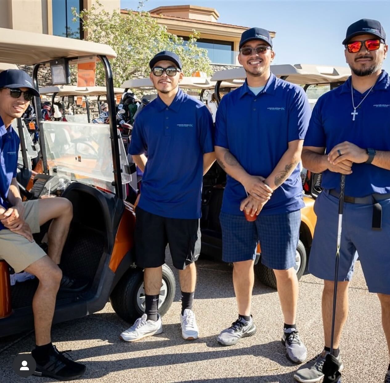 Four men in blue shirts and caps pose by golf carts in an outdoor setting.