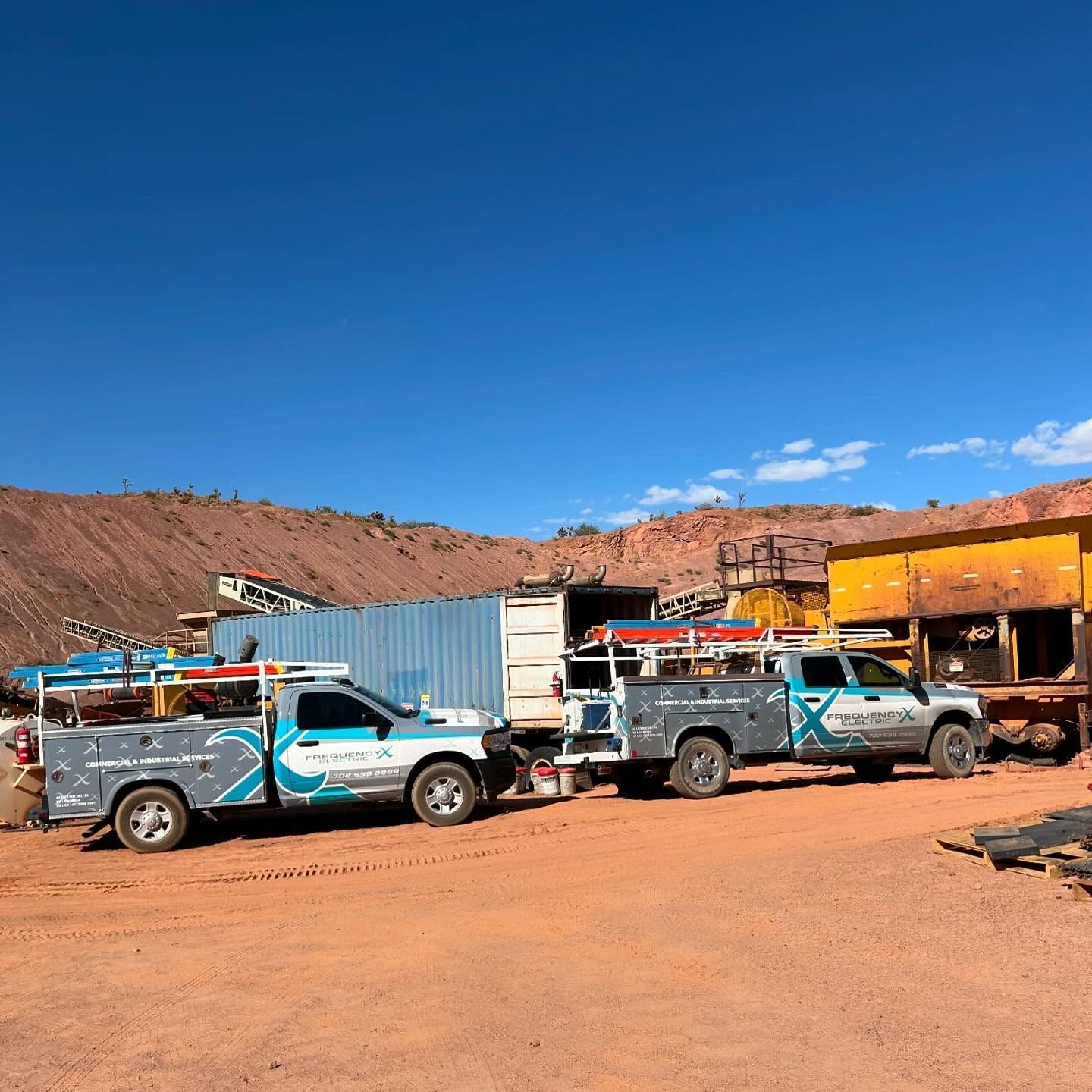 Two work trucks parked in a dusty lot near red rock formations under a blue sky.