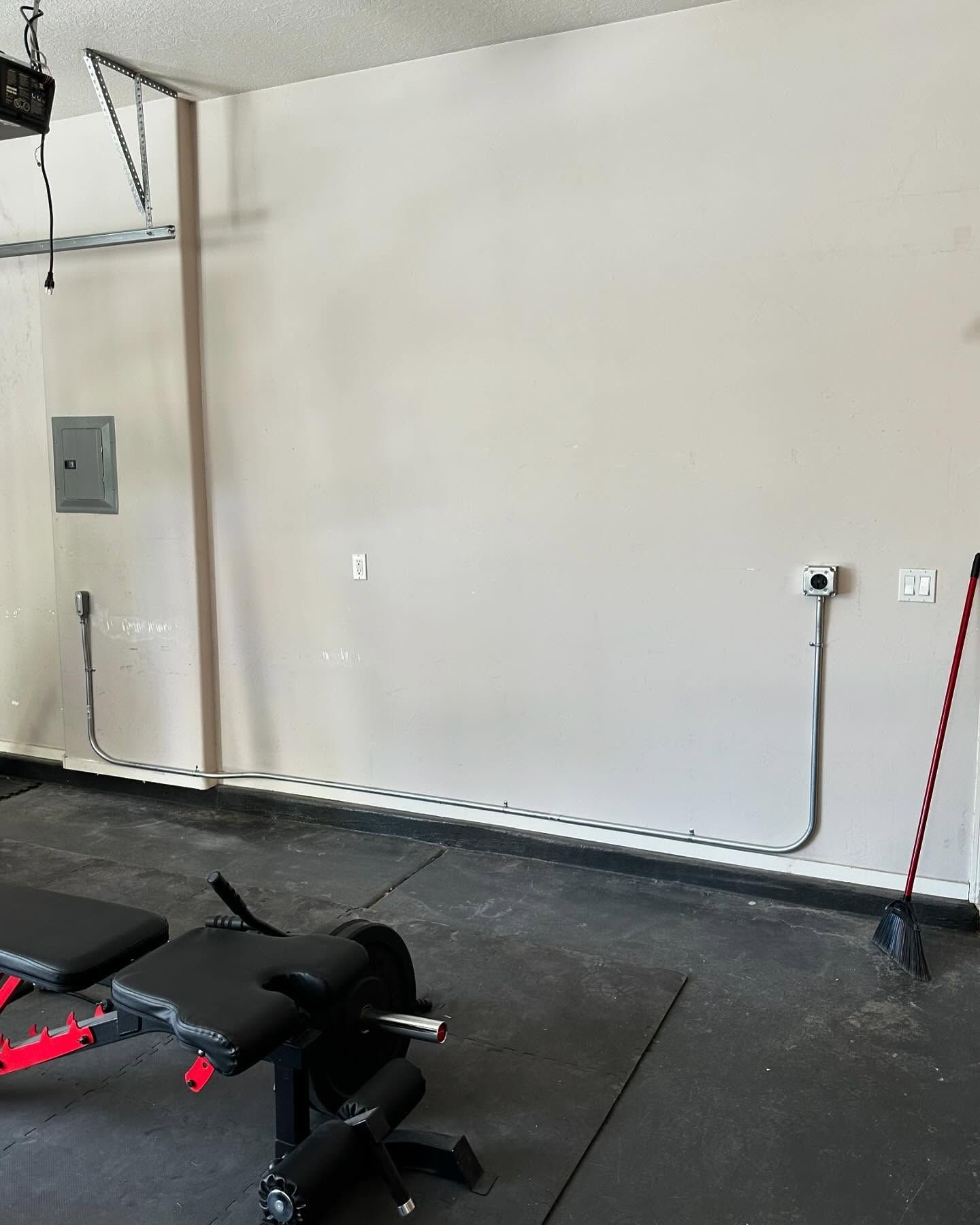 Garage gym with weight bench, electrical boxes, exposed conduit, and a broom.