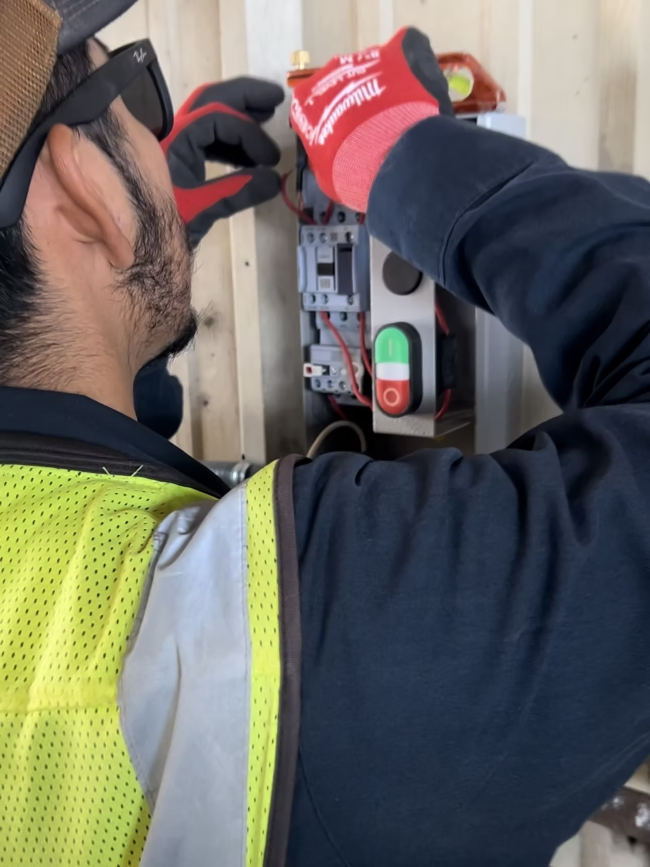 Electrician wiring electrical panel; red and black gloves, safety vest.