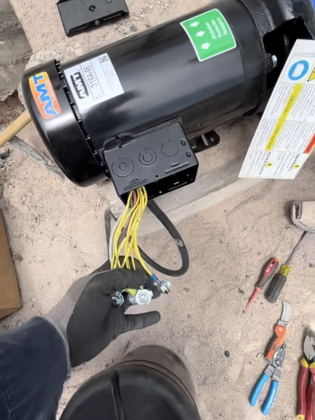 A person wiring a black electric motor with yellow wires, tools visible, on concrete.