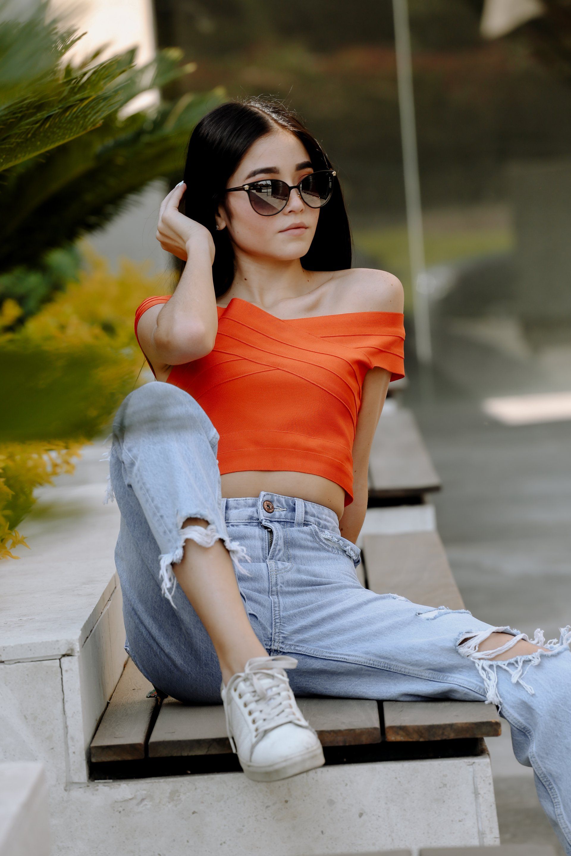 Mujer con top naranja y jeans rotos, con gafas de sol, sentada en un banco al aire libre.Retrato profesional cinematográfico para marca personal y especialistas médicos realizado por RomRom.VERSACHE