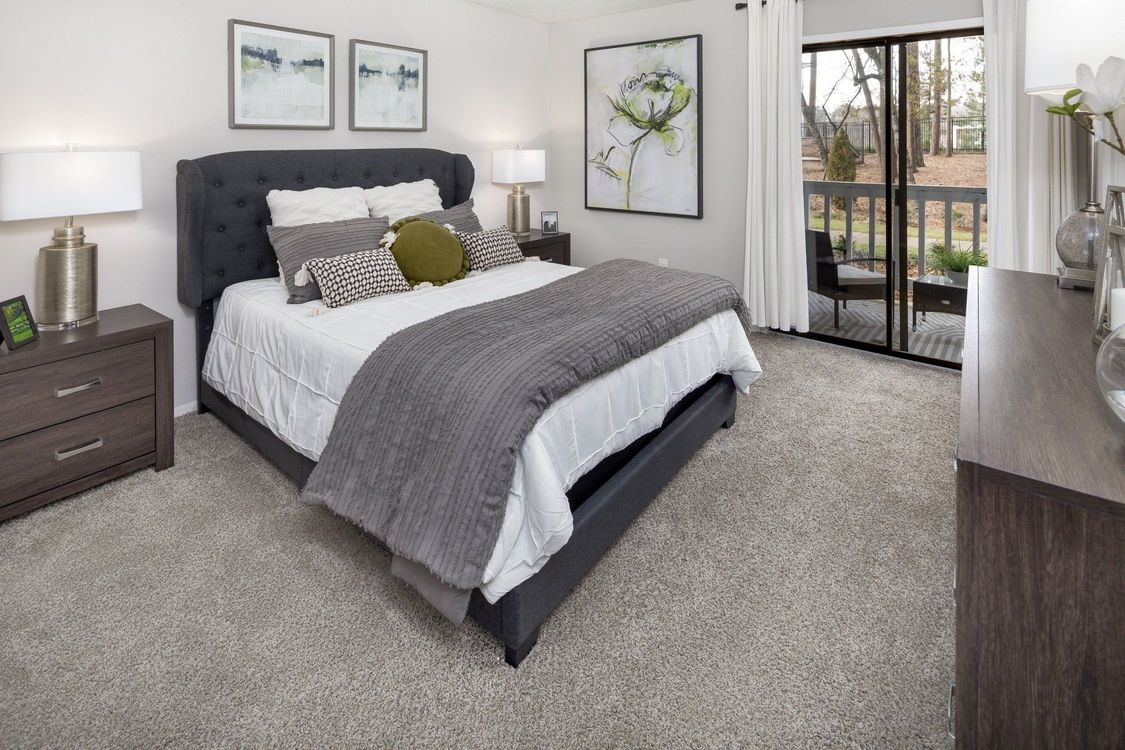 Bedroom with gray carpet, bed, and balcony access. Neutral tones, artwork, and bedside tables.