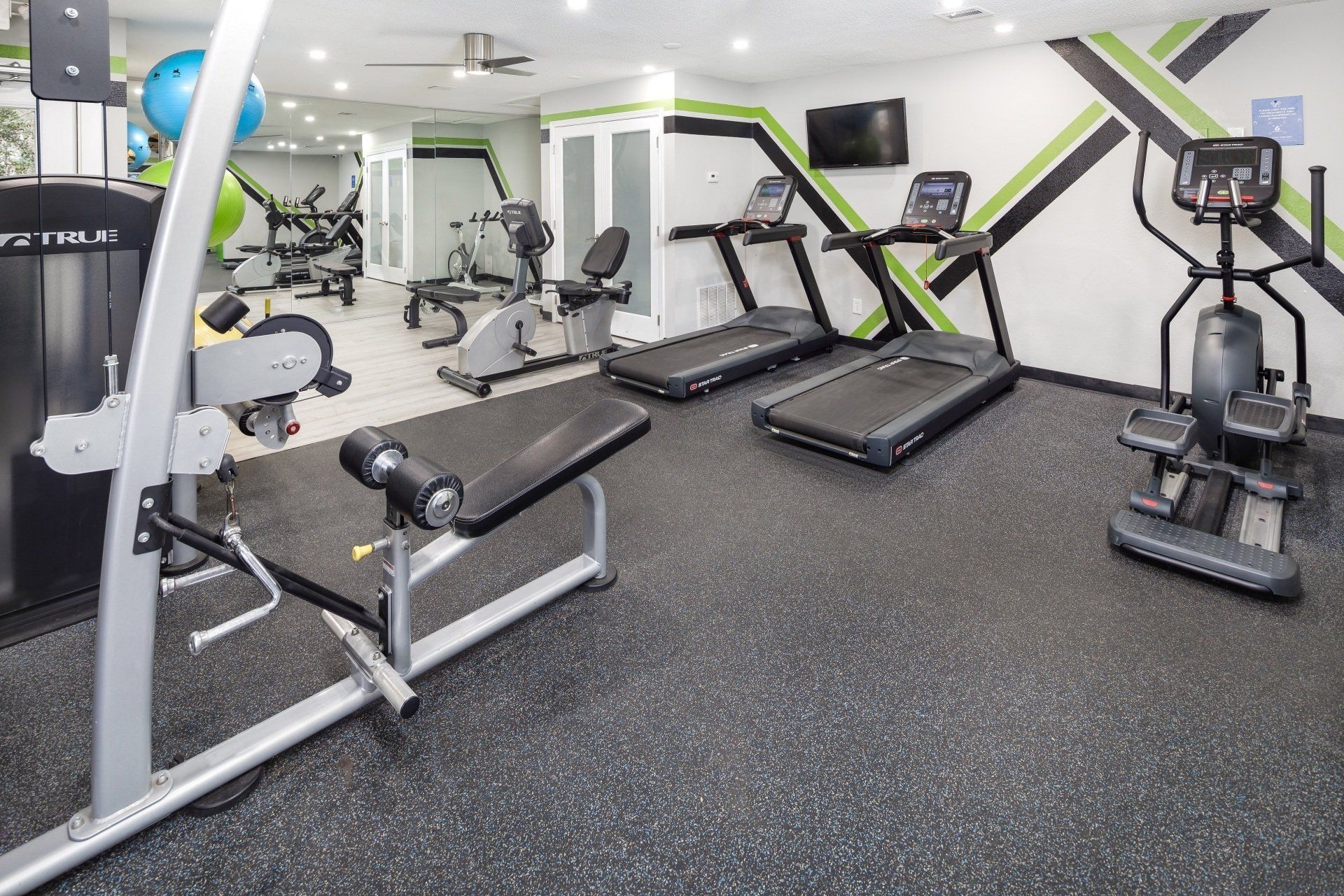 Gym with treadmills, elliptical, weight machines, and exercise balls on a black rubber floor; light green and black accents on walls.