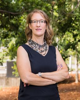 Arien Muzacz, Ph.D. wearing glasses and a black tank top is standing with her arms crossed .