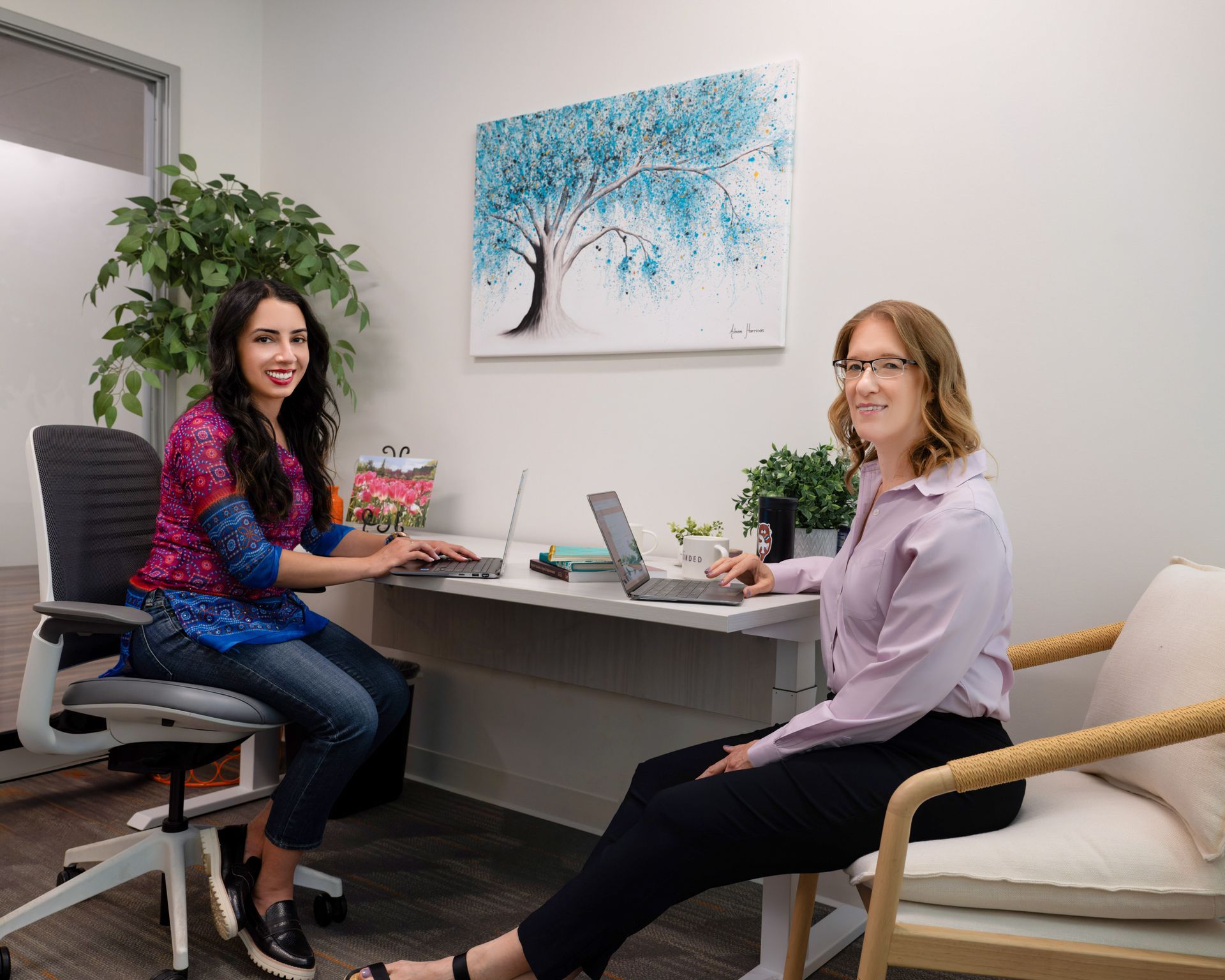 Two women are sitting at a desk with laptops.