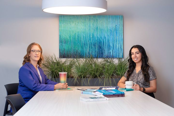 Anjabeen and Arien are sitting at a table in front of a painting.