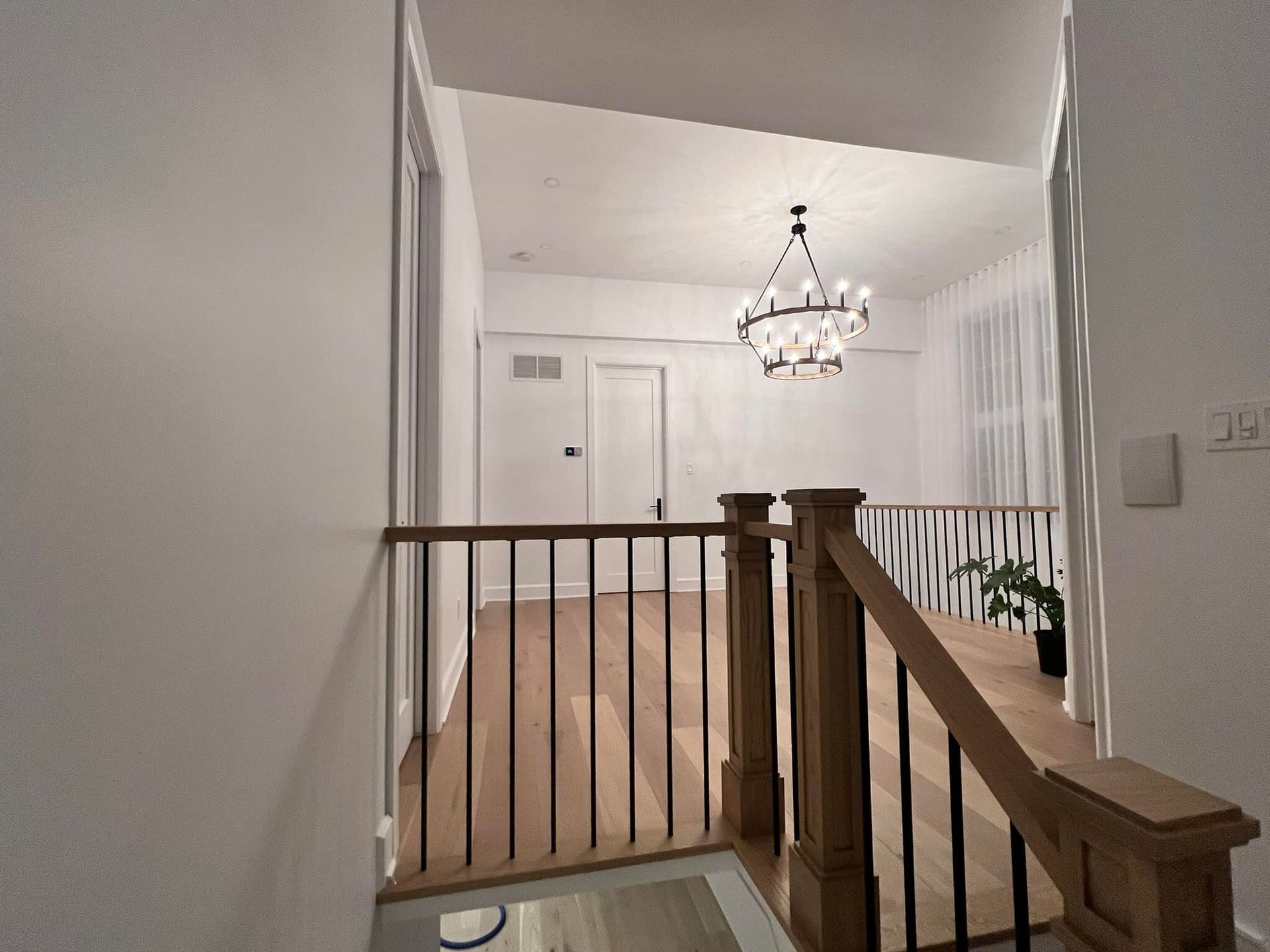 Vue depuis un palier d'un couloir avec parquet, rampe sombre et lustre. Deux portes et des murs blancs se dessinent au loin.