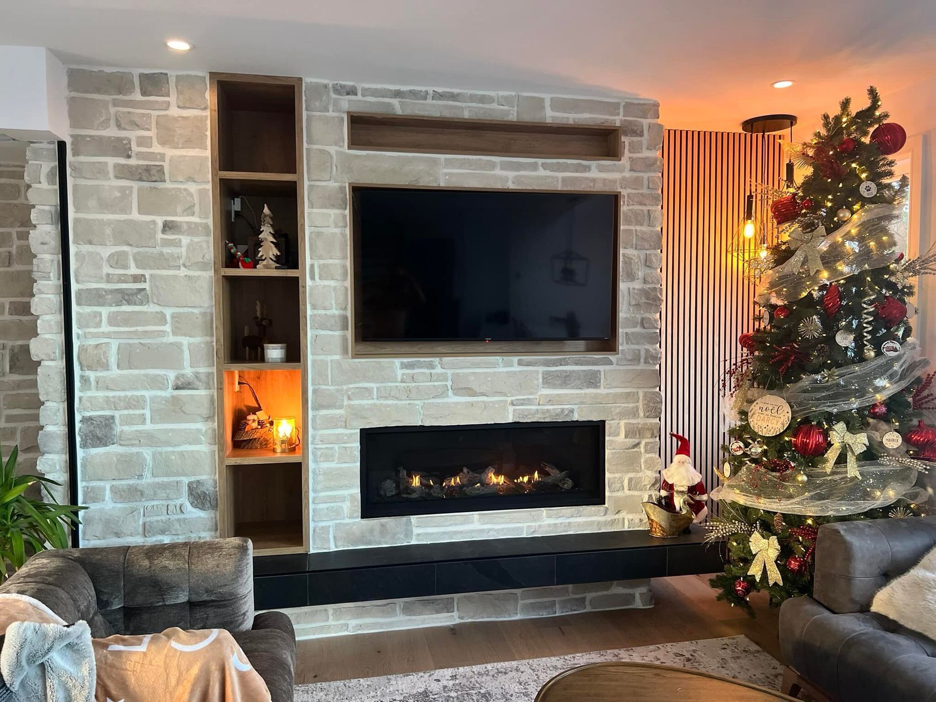 Salon avec cheminée en pierre, étagères encastrées et sapin de Noël décoré de lumières et de décorations. Un téléviseur est installé au-dessus de la cheminée.